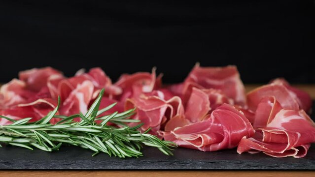 Prosciutto crudo or jamon meat with rosemary on serving board on black background. Sliced into thin pieces of ham puts on cutting board. Put jamon on serving board. Dish serving, food serving process