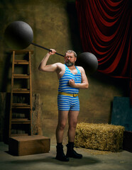 Retro circus. Cinematic portrait of retro circus strongman wearing striped sports swimsuit holding barbell over dark circus backstage background. Art, fashion, style