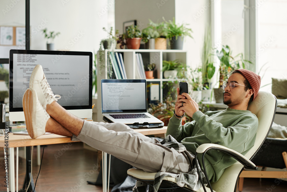 Poster Young restful IT engineer sitting in armchair by workplace with computer monitor and laptop snd texting in smartphone at break