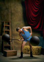 Vintage portrait of retro circus strongman wearing blue striped sports swimsuit training with barbell over dark circus backstage background. Art, fashion, style © master1305