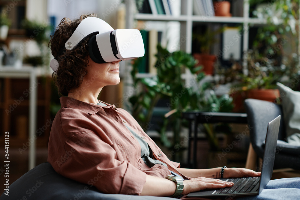 Fototapete Side view of young female programmer in vr headset using laptop to control work with virtual stuff while sitting in openspace office