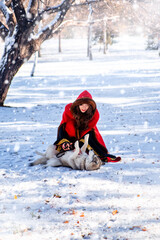 Fototapeta na wymiar A young woman in a red hooded raincoat with a gray husky dog on the background of a winter forest and snowfall.