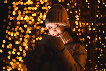 Girl and gift box with Christmas lights. Smiling girl in a hat, scarf looking at the camera at night with blurry lights. Holidays theme