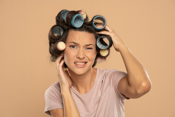 Beautiful nervous woman with hair curlers scratching her head on a beige background