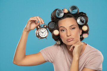 Beautiful bored woman with hair curlers holding a clock on a blue background