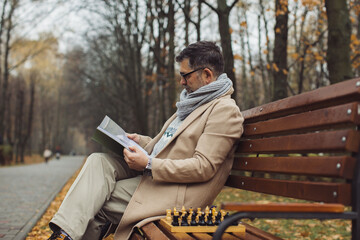 Fototapeta premium An elderly man reads a magazine while sitting on a bench in an autumn park. A man is waiting for his friend to play chess.