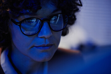 Fototapeta na wymiar Selective focus of face of serious businesswoman in eyeglasses sitting in front of computer late at night in office and working overtime