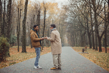 Friends, a senior and a young man walking and talking and drinking coffee together in the autumn park..
