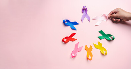 Hand holding pink ribbon which is among colorful ribbons on pink background, cancer awareness,...