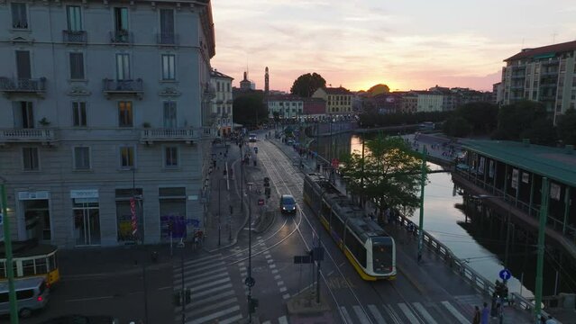 Forwards tracking modern tram unit passing through crossroads and driving along water. People enjoying sunset time in city. Milano, Italy