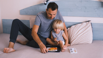 A small caucasian boy with his father on the bed draw with colored pencils.