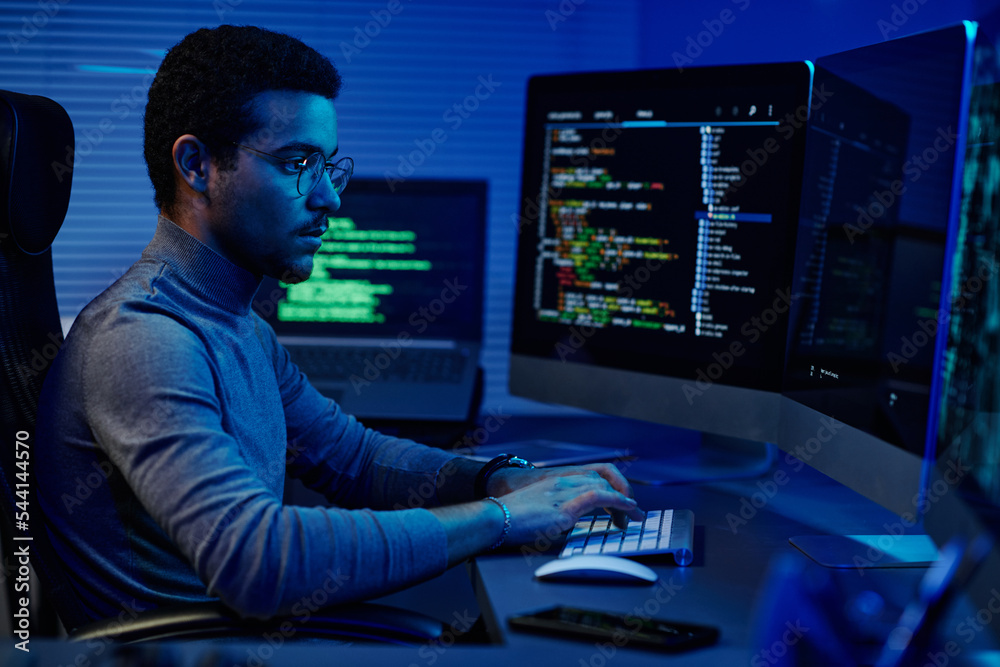 Poster Young serious software developer typing on desktop keyboard while working in front of computer monitors in server room