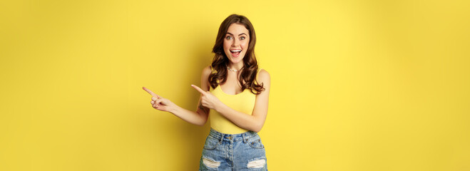 Beautiful young woman pointing fingers at banner, logo or advertisement, smiling amazed, standing in summer clothes, yellow background