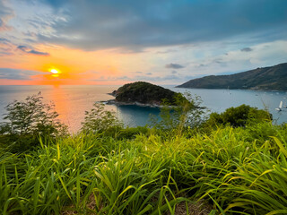 Summer island sunset background. Beautiful tropical landscape aerial view. Colorful sky with orange sun over green grass meadow. Turquoise sea, yachts, ocean bay. Windmill Viewpoint, Phuket, Thailand