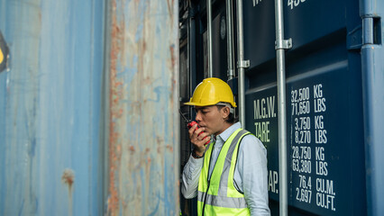 Young Asian man in vest and hardhat looking upwards towards container shipment and sharing details and information while talking to coworker using walkie-talkie radio device