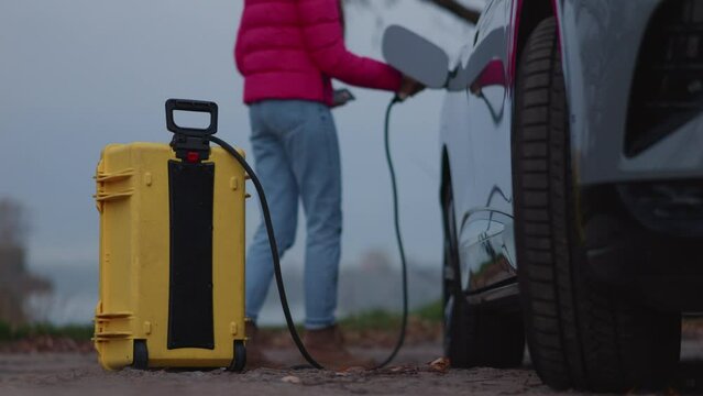 Girl uses home-invented portable battery to charge electric car out of the city,turns it on with phone,takes photos of nearest lake. Unrecognisible woman,pink jacket,foggy day.High quality 4k footage