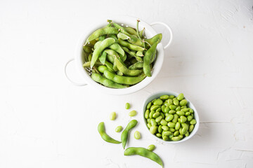 Aerial view of pods in drainer and edamame grains in white bowl, on white table, horizontal, with copy space