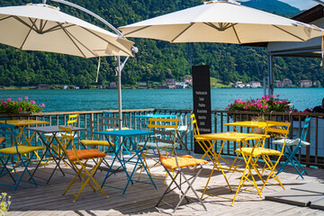 Beautiful terrace of modern cafe with bright colorful chairs on the shore of Lake Lugano in Morcote, Switzerland, with splendid views of the mountains and the lake on a sunny summer day.