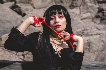 Young, sexy and vampiric hispanic goth girl with black dress seated and playing with "blood" (red ribbon) in a wood bench between big rocks under sunlight