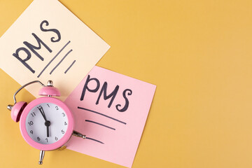 Pink mechanical alarm clock and the inscription on the sheets of paper "PMS" on a yellow background. The concept of the time of critical days, women's health. Space for text.