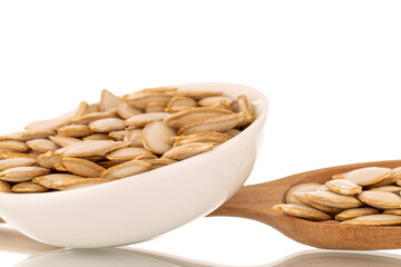 Unpeeled raw pumpkin seeds in a white ceramic saucer with a wooden spoon, macro, isolated on white background.