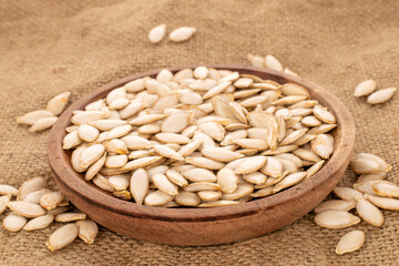 Unpeeled raw pumpkin seeds in a wooden plate on a jute cloth, macro.