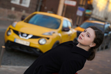 Fototapeta na wymiar beautiful girl in the city. person in the street. a cute teenager in a black coat against a background of a blurred yellow car.