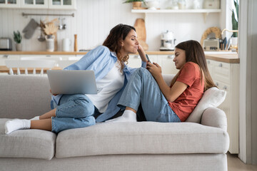 Naklejka na ściany i meble Young smiling woman sitting on sofa with laptop spying on child teen girl using cellphone, mom and kid spending leisure time together on devices. Parental control and smartphone generation