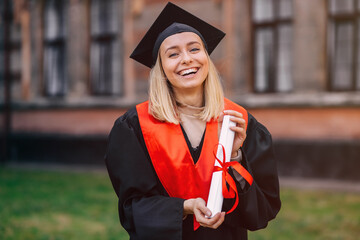 A happy graduate stands in a university garden in a mantle with a diploma in her hand, smiles and...