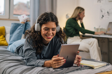 teenager caucasian female with headphones using digital tablet while laying on bed in dorm room