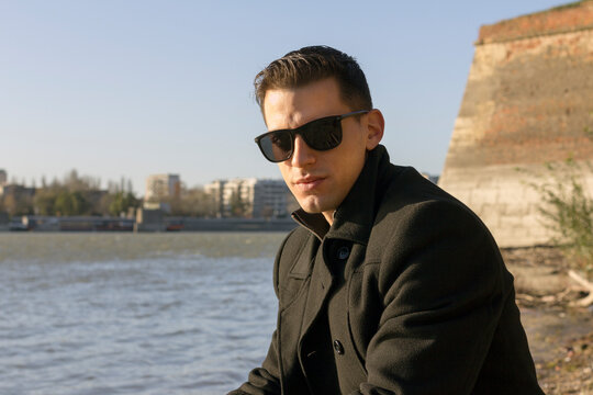 Stock photo of a handsome guy sitting by the river looking at the camera with sunglasses.