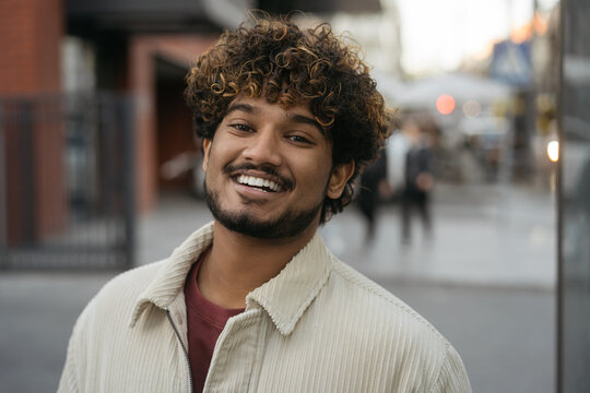 Smiling stylish Indian man wearing casual clothing looking at camera on the street. Happy asian fashion model with curly hair posing for picture outdoors  