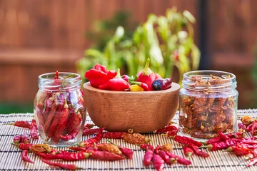 Fotobehang Still life picture of two jars with dried red and yellow chilli pepers with small wooden bowl with freshly harvested diferent kinds of red chilli peppers. Home garden harvest of spices in summer time. © jdmfoto