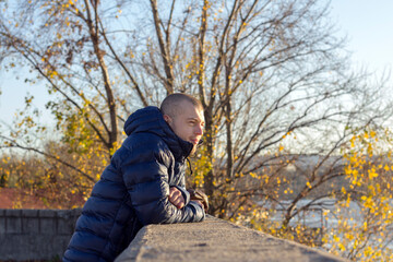Young Man leaning on the fence and looking at the river, relaxing on a winter sunny day.
