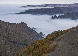 Mountain hills with yellow trees and scree and rocky protruding from above from the clouds that creep down the valley, autumn landscape in the Caucasus mountains