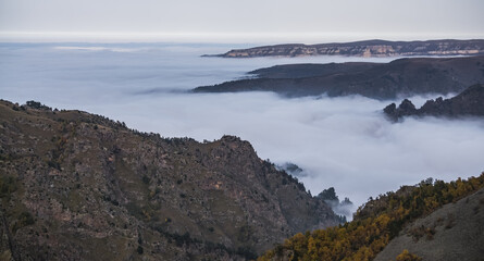 Rocky vertical cliffs of mountains protruding from above above the fog and clouds on an autumn day in the mountains of the Caucasus, hills with scree and yellowed vegetation