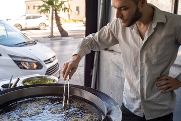 Fototapeta premium Young White Man in Grey Shirt Moves a Clamp and Fries Falafel Balls in a Giant Pan Filled with Boiling Oil in Middle Eastern Restaurant