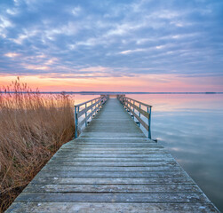 Calm Lake at Sunrise with Long Wooden Pier and Reeds under cloudy sky at sunrise