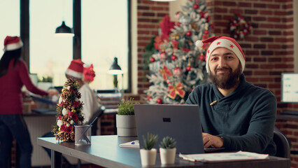 Portrait of project manager with santa hat working on laptop during christmas holiday season. Using...