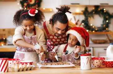 Happy ethnic family father and two kids in festive outfit making Christmas cookies together in...