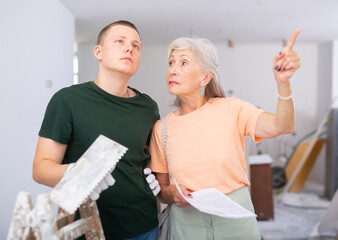 Young man with floated trowel discussing repair works in apartment with senior woman, talking about...