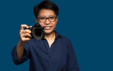 A woman photographer with short black hair holds a digital camera while standing on a blue background