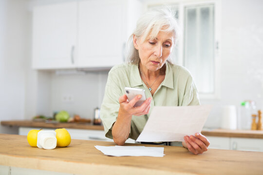 Worried Eldelry Woman Reading Papers And Having Phone Conversation At Kitchen At Home