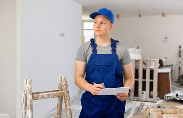 Teenage boy in blue overall checking documents in construction site during workday on part-time job.