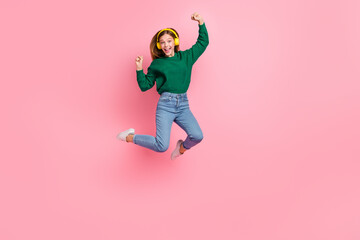 Full length photo of shiny lucky young lady wear green sweater jumping high listen music rising fists isolated pink color background