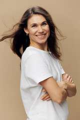 Fototapeta premium portrait of a cute, happy, beautiful brunette woman, with hair blowing in the wind, in a white tank top, standing sideways with her arms crossed on her chest, and smiling at the camera