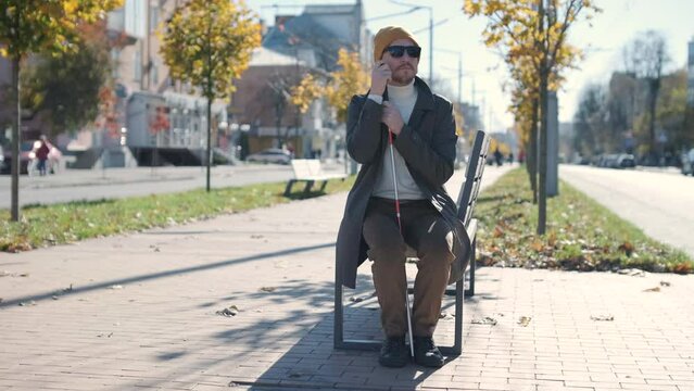 Young blind man with smartphone sitting on bench in park in city, calling