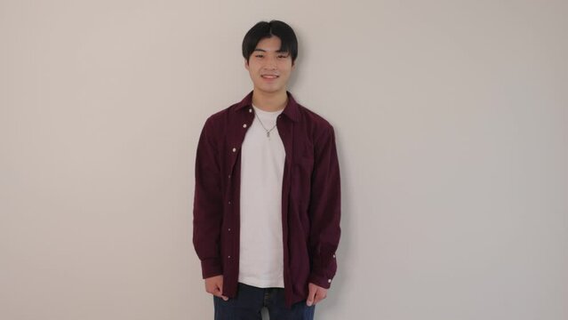 Asian boy standing still against the white wall and smiles. Wide shot.