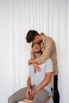 Gay couple hugging from behind and kissing on forehead with white curtain background