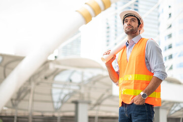 Young caucasian man holding a big paper, a guy wearing a light blue shirt and jeans with an orange vest and white helmet for security in the construction area.